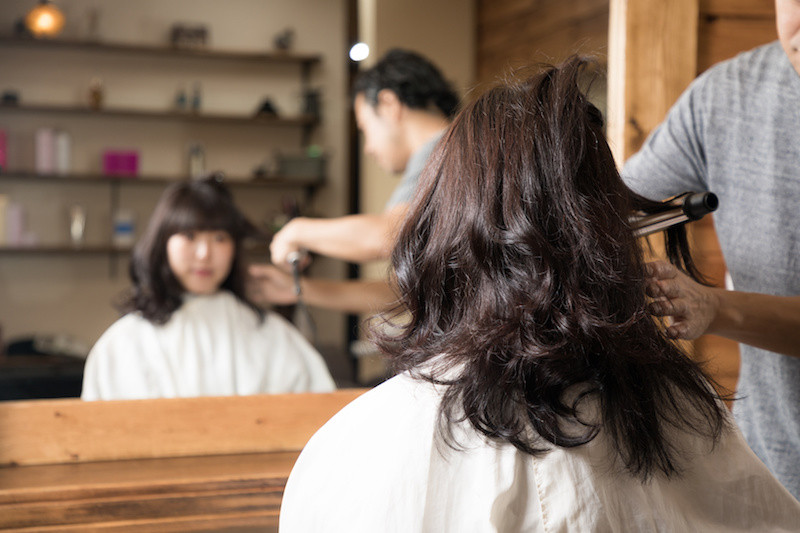美容師に髪の毛をコテでカールしてもらっている茶髪ロングヘアの女性の後ろ姿と鏡に映っている姿